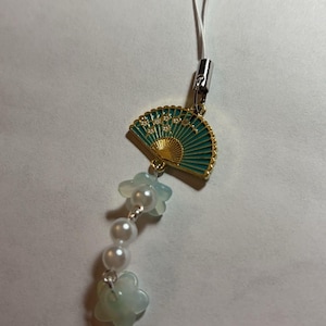 May include: A teal and gold fan charm with white pearl beads and white flower charms attached to a white string.
