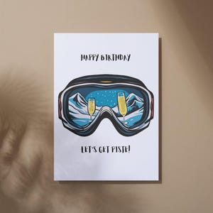 May include: A white card with the text "Happy Birthday" and "Let's get piste!"  A pair of blue ski goggles with a snowy mountain scene reflected in the lenses. Two champagne glasses are visible in the reflection.