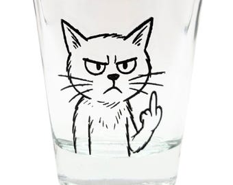May include: Clear shot glass with a black and white illustration of an angry cat giving the middle finger. The cat has a grumpy expression with furrowed brows and a frown. The glass is cylindrical with a thick base.