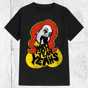 May include: A black short-sleeved t-shirt featuring a graphic print of a stylised face with red hair and black liquid dripping from the mouth. Below the face, the text YEAH YEAH YEAHS is printed in bold black letters against a yellow background.