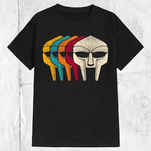 以下が含まれることがあります： 黒いTシャツ。黄色、ターコイズ、オレンジ、赤、白のマスクのレトロなグラフィック。デザインはヴィンテージ感があります。シャツはクラシックなクルーネックです。