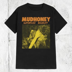 May include: A black short-sleeved t-shirt featuring the orange text MUDHONEY and SUPERFUZZ BIGMUFF above a yellow and black graphic illustration of a person playing an electric guitar in a dynamic, high-energy performance pose.