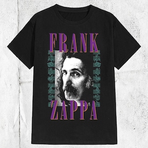 Puede incluir: Camiseta negra con un retrato de Frank Zappa y el nombre "FRANK ZAPPA" en letras moradas y verdes. El diseño incluye notas musicales alrededor del retrato. La camiseta es de cuello redondo.