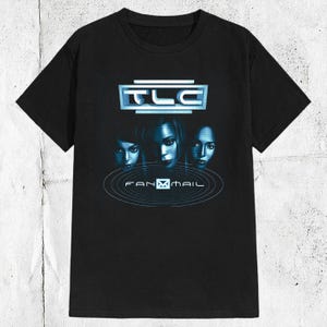 May include: A black short-sleeved t-shirt featuring a blue graphic print on the chest. The design includes the text TLC at the top, a central image of three faces, and the text FANMAIL below, all surrounded by concentric circular line patterns.