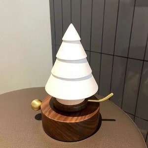 Op de afbeelding: Een witte keramische kerstboom muziekdoos met een houten basis en een gouden opwindsleutel. De muziekdoos staat op een bruin lederen oppervlak.