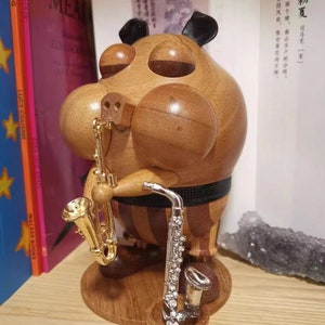May include: A wooden figurine of a pig playing a saxophone. The pig is wearing a black and white striped shirt and has a black and white striped hat. The pig is holding a gold saxophone in its right hand and a silver saxophone in its left hand. The pig is standing on a wooden base.