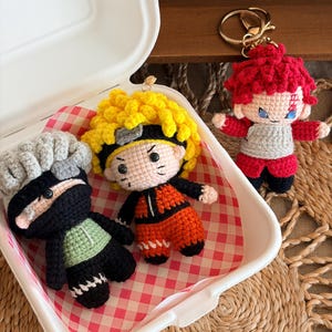May include: Three small crocheted character keychains. One has grey hair and a black mask, one has spiky yellow hair and an orange outfit, and one has red hair with a symbol on the forehead and a red and grey outfit. They are made of yarn.
