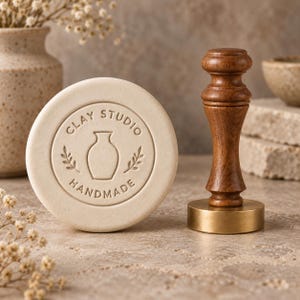 Può includere: Un disco circolare in argilla color bianco sporco con il testo in rilievo CLAY STUDIO HANDMADE e un'icona centrale a forma di vaso con foglie, accanto a un timbro in legno con base in ottone e manico tornito per imprimere l'argilla.