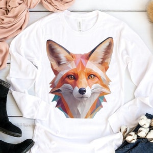 Puede incluir: Camiseta blanca de manga larga con un diseño de zorro geométrico en tonos naranja, rojo y azul. La camiseta está colocada sobre una superficie de madera blanca con una bufanda, unos vaqueros y una boina.