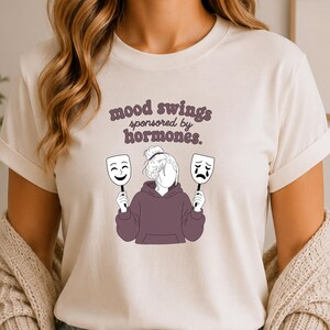 Può includere: Una t-shirt a maniche corte color crema con la grafica di una persona in felpa con cappuccio viola scuro che tiene in mano due maschere, una sorridente e una piangente. Il testo sopra la grafica recita mood swings sponsored by hormones.