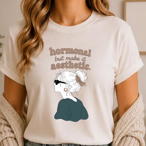 Könnte beinhalten: Ein cremefarbenes Kurzarm-T-Shirt mit einer grafischen Illustration einer Person mit Dutt und Sonnenbrille. Der Text "hormonal but make it aesthetic." ist in Braun über der Illustration aufgedruckt. Das T-Shirt wird mit einer blauen Jeans kombiniert.