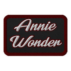 Könnte beinhalten: Ein gestickter schwarzer und roter Aufnäher mit dem Namen "Annie Wonden" in weißer Kursivschrift.