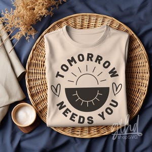 May include: A beige t-shirt with a black graphic design that says "Tomorrow Needs You" with a sun and hearts.