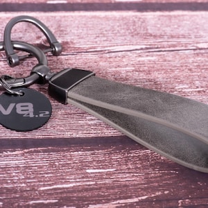 May include: Gray leather keychain with a metal clasp and a round black tag. The tag has the text "V8 4.2" in silver. The keychain has a loop for attaching keys and a gray leather strap. The background is a wooden surface.