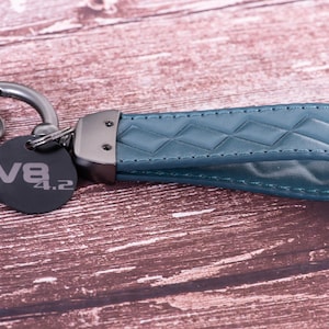 Può includere: Portachiavi in pelle color teal con cinturino trapuntato a diamante e chiusura in metallo. È attaccato un tag circolare nero con la scritta "V8 4.2". Il portachiavi è progettato per chiavi di auto.
