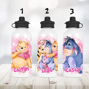 May include: Three white water bottles with black lids, each featuring a different Winnie the Pooh character. The bottles have pink watercolour backgrounds and personalized names: Caitlin, Leila, and Casey. The bottles are on a light wood surface.