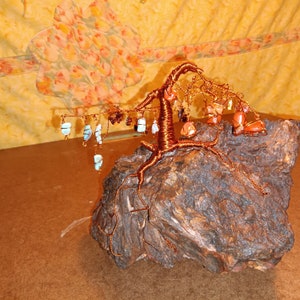 Puede incluir: Una escultura de árbol de alambre de cobre con varias piedras preciosas de colores colgando de las ramas. El árbol está enraizado en un trozo de madera áspera de color marrón.