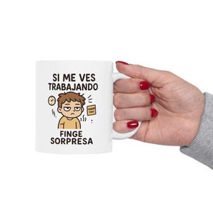 May include: White ceramic mug with the text "SI ME VES TRABAJANDO FINGE SORPRESA" and a cartoon illustration of a stressed person. The mug is held by a hand with red nail polish.