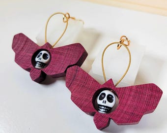 May include: A pair of earrings featuring a wooden bat design with a skull detail. The bats are a deep burgundy colour and the skulls are white with black details. The earrings are suspended from gold hoops.