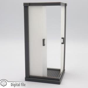 Może przedstawiać: Szary plastikowy model kabiny prysznicowej ze  slidowymi drzwiami. Drzwi są półprzezroczyste i mają po jednej małej klamce z każdej strony. Kabina prysznicowa ma szarą podstawę z fakturowaną powierzchnią.