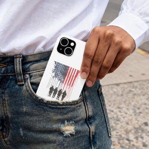 May include: White phone case featuring an American flag design with a silhouette of soldiers. The flag is depicted with red and white stripes and a blue field of stars. The case is in a pocket.