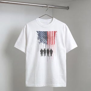 May include: White t-shirt featuring a watercolor design of the American flag with a silhouette of soldiers. The flag design includes red and white stripes, and a blue field with white stars. The shirt is hanging on a metal rod.
