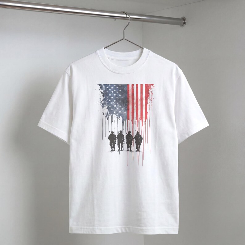 May include: White t-shirt featuring a watercolor design of the American flag with a silhouette of soldiers. The flag design includes red and white stripes, and a blue field with white stars. The shirt is hanging on a metal rod.