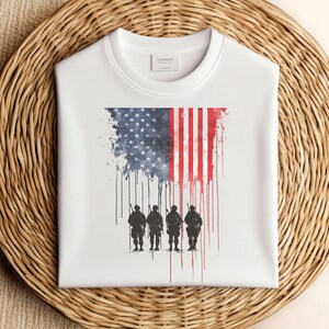 May include: White sweatshirt featuring an American flag design with a distressed effect. The flag transitions into a silhouette of four soldiers. The word "SALESON" is printed on the label. The sweatshirt is displayed on a woven surface.