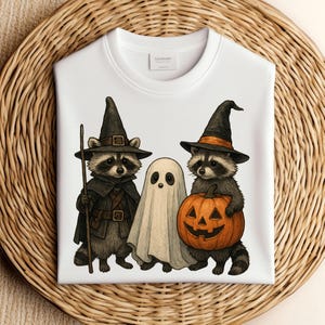 Może przedstawiać: Biała bluza z nadrukiem na Halloween. Przedstawia dwa szopy w kapeluszach czarownic i ducha. Jeden szop trzyma laskę, drugi rzeźbioną dynię. Projekt jest w stylu kreskówkowym.