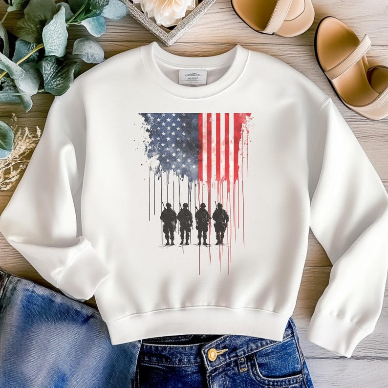 May include: White crew neck sweatshirt featuring a distressed American flag design with a silhouette of four soldiers standing at attention. The flag design incorporates red, white, and blue colors, with a dripping effect. A patriotic themed apparel item.
