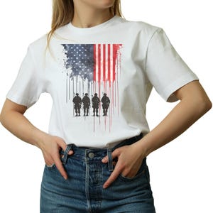 May include: White t-shirt featuring a graphic of the American flag with a distressed effect and silhouettes of soldiers. The flag design incorporates the stars and stripes, with a dripping paint effect. The shirt is a classic crew neck style.