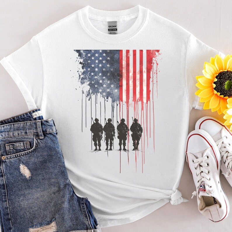 May include: White t-shirt featuring an American flag design with a watercolor effect. The flag's stars and stripes are depicted with a dripping effect, and silhouettes of soldiers stand below. The shirt is paired with denim shorts and white sneakers.