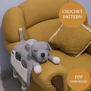 Può includere: Un organizer per divano a forma di cane all'uncinetto grigio con dettagli bianchi, un portabicchieri e una tasca. Il cane è sdraiato su una poltrona gialla. L'immagine include il testo "CROCHET PATTERN" e "PDF DOWNLOAD".