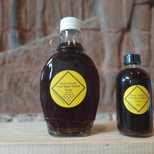 Puede incluir: Dos botellas de vidrio de Door County Pure Black Walnut Syrup. La botella más grande tiene una tapa blanca y un asa, y la botella más pequeña tiene una tapa negra. Ambas botellas tienen etiquetas amarillas con texto negro. El jarabe es marrón oscuro.