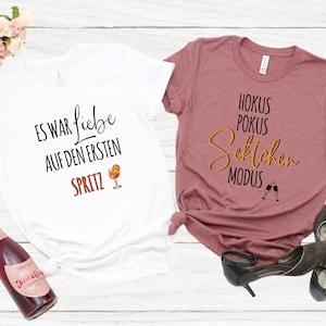 Può includere: Due t-shirt con testo e un'immagine di un bicchiere di vino. La t-shirt bianca dice "Es war Liebe auf den ersten Spritz" e la t-shirt rosa dice "Hokus Pokus Sektchen Modus".