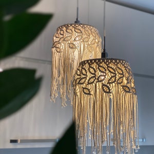 May include: Two macrame pendant lights in a white, cream and beige colour scheme. The lights are hanging from the ceiling and are illuminated. The lights are made of a woven rope material and have a fringe design.