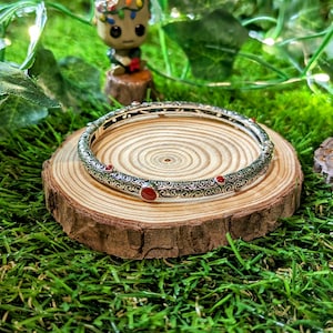 May include: A silver bangle bracelet with an intricate pattern and red gemstone details is displayed on a wooden slice. The bracelet is set against a backdrop of green grass and decorative foliage.