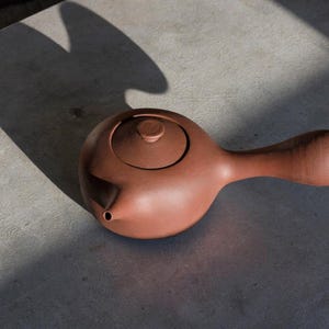 May include: A reddish-brown ceramic teapot with a rounded body and a long, curved handle. The teapot has a small spout and a lid with a knob. The teapot is sitting on a gray surface, with a shadow cast across it.
