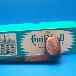Pode incluir: Um cachimbo de madeira marrom com uma haste preta e uma caixa verde com o texto "The Guildhall London Made in England" nela.