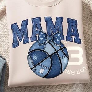 以下が含まれることがあります： 薄いピンク色のスウェットシャツ。青いデニムの文字で「MAMA」と書かれています。青いデニムのバスケットボールにリボンがついており、その下に配置されています。「3」の数字がバスケットボールの右側に表示されています。