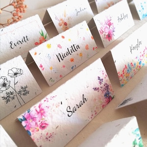 Może przedstawiać: Złożone karty z nazwiskami takimi jak "Everett", "Julia" i "Ashley". Karty zdobią kolorowe kwiatowe wzory. Papier ma teksturowany, ręcznie robiony wygląd. Nazwiska są napisane stylową czcionką.