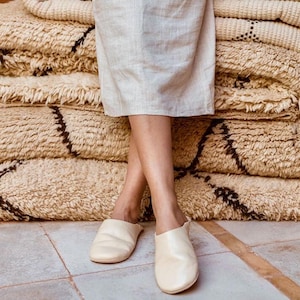 Puede incluir: Un par de zapatillas de cuero de color blanco roto con punta puntiaguda. Las zapatillas son usadas por una persona de pie sobre una pila de alfombras beige y marrones.