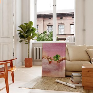 May include: A pink and white canvas print featuring a painting of two hands reaching towards each other. One hand is holding a cup of liquid and the other hand is holding a sprig of green leaves. The painting is set against a background of clouds.