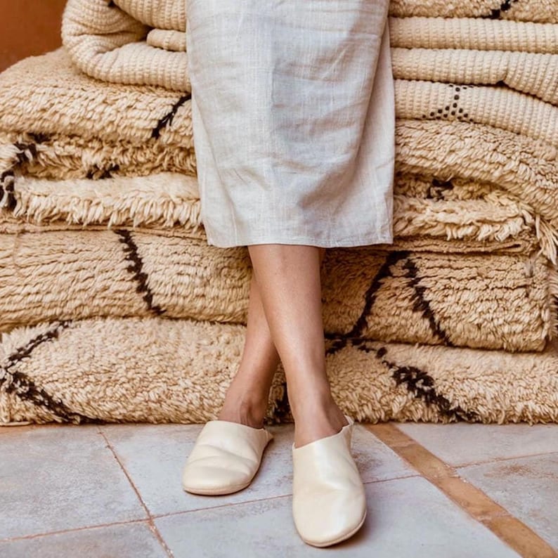Puede incluir: Un par de zapatillas de cuero de color blanco roto con punta puntiaguda. Las zapatillas son usadas por una persona de pie sobre una pila de alfombras beige y marrones.