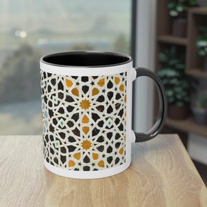 Peut inclure: Mug en céramique blanche avec une bordure et une poignée noires. La tasse présente un motif géométrique dans des tons de blanc, noir et or.