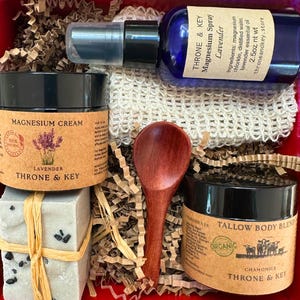 May include: A red gift box filled with self-care products. It includes a blue spray bottle, two jars with black lids, a wooden spoon, and two bars of soap tied with twine. The products are labeled with the brand "Throne & Key."