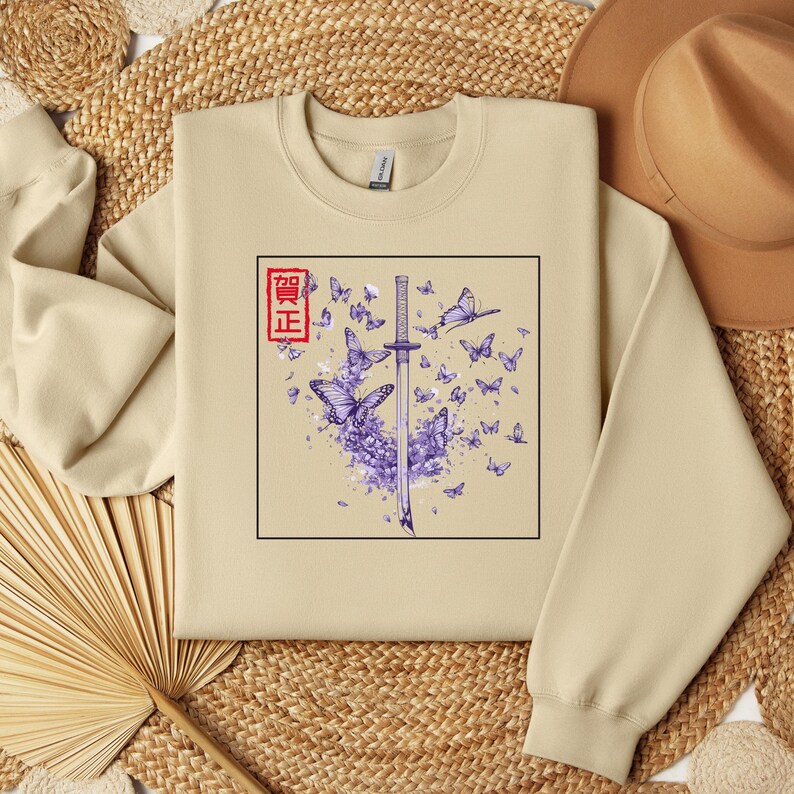 May include: A beige sweatshirt with a graphic print of a purple sword and butterflies. The Japanese word "真正" is printed in red on the graphic.