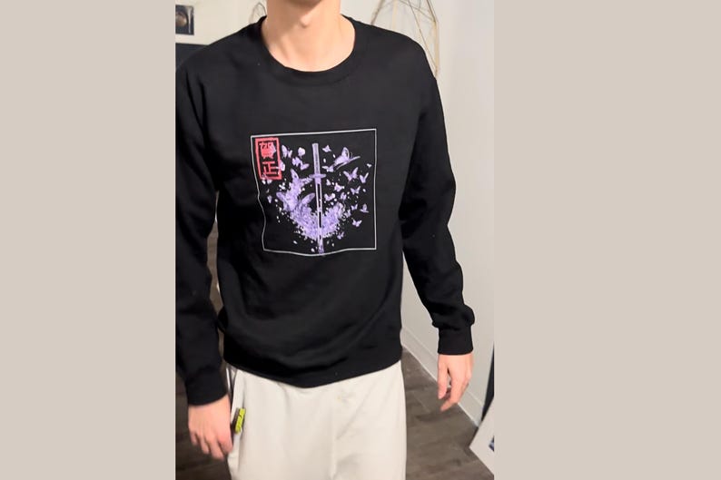 May include: Black long-sleeve sweatshirt with a graphic print of a sword surrounded by purple butterflies. The graphic also includes the text "悟" in red.