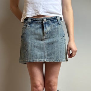 May include: A light-wash denim skirt with scattered dark embellishments. The skirt has a classic five-pocket design and a button closure. The model is wearing the skirt with a white top and dark brown knee-high boots.