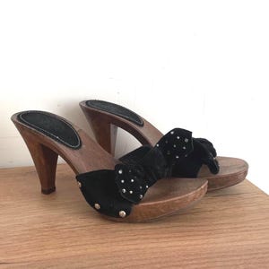 May include: A pair of black suede mules featuring a wooden platform sole and a high wooden heel. The black suede upper is adorned with a bow detail and small silver-toned metal studs. The footbed is lined with black material and stitched edges.